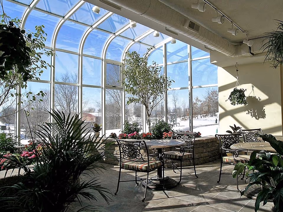 Sunlit indoor seating area with round tables and wrought iron chairs with striped cushions, surrounded by various green plants and flowers. Large curved glass windows provide a view of a snowy outdoor landscape with trees and buildings in the distance.