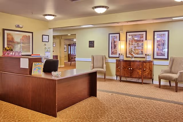 Reception area with a wooden desk, a black office chair, and a 'Welcome' sign. The room has beige walls with framed artwork, two striped armchairs, a wooden sideboard with decorative lamps, and carpeted flooring.