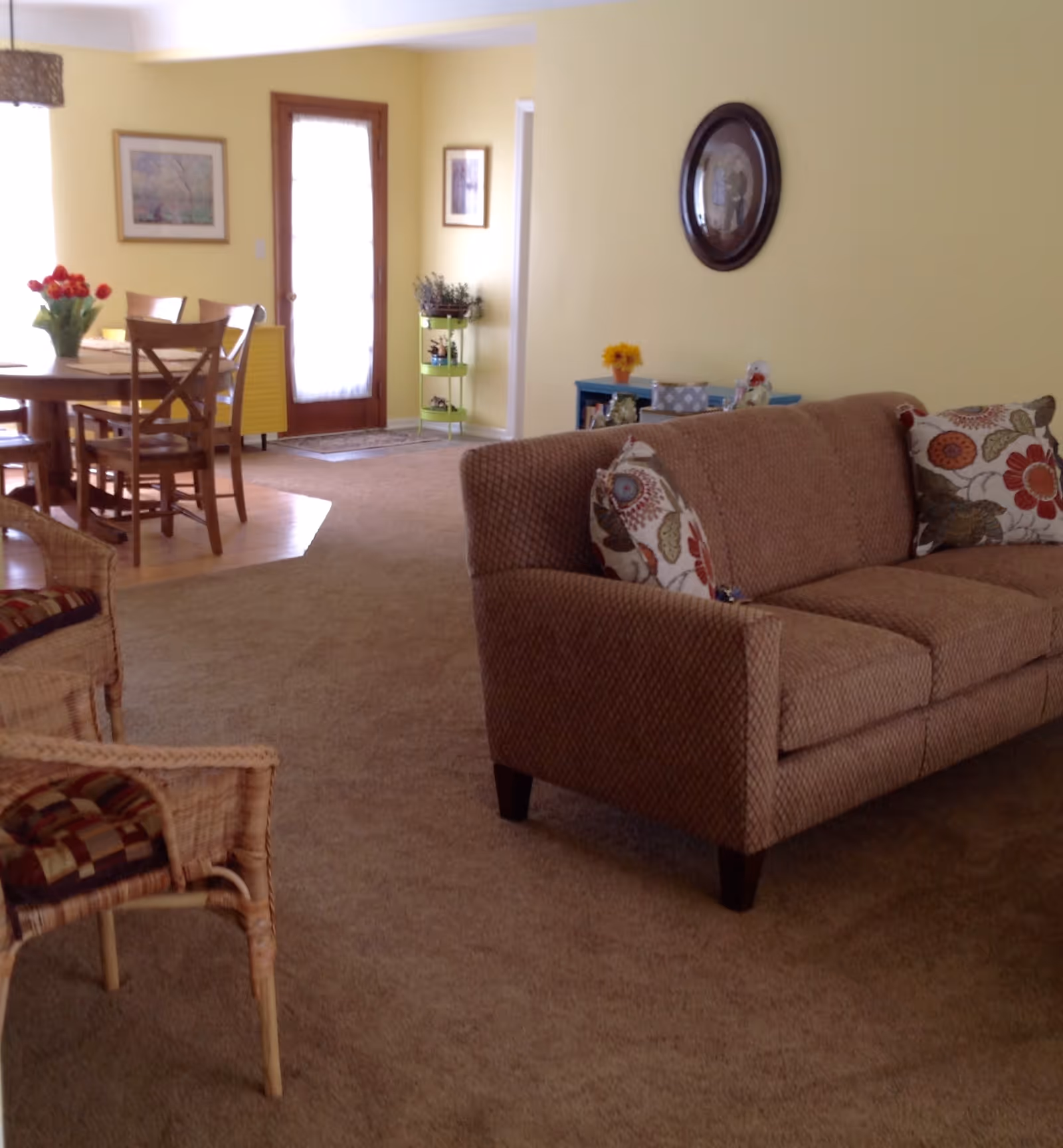 A cozy living room area with a brown upholstered sofa adorned with floral patterned pillows. To the left, there are wicker chairs with cushions and a wooden dining table with chairs. The walls are painted yellow, and there is a wooden door with a window letting in natural light. The room has a carpeted floor with a small section of hardwood near the dining area. Decorative items include framed pictures on the walls, a small green shelf with plants, and a blue cabinet with flowers and other decor.