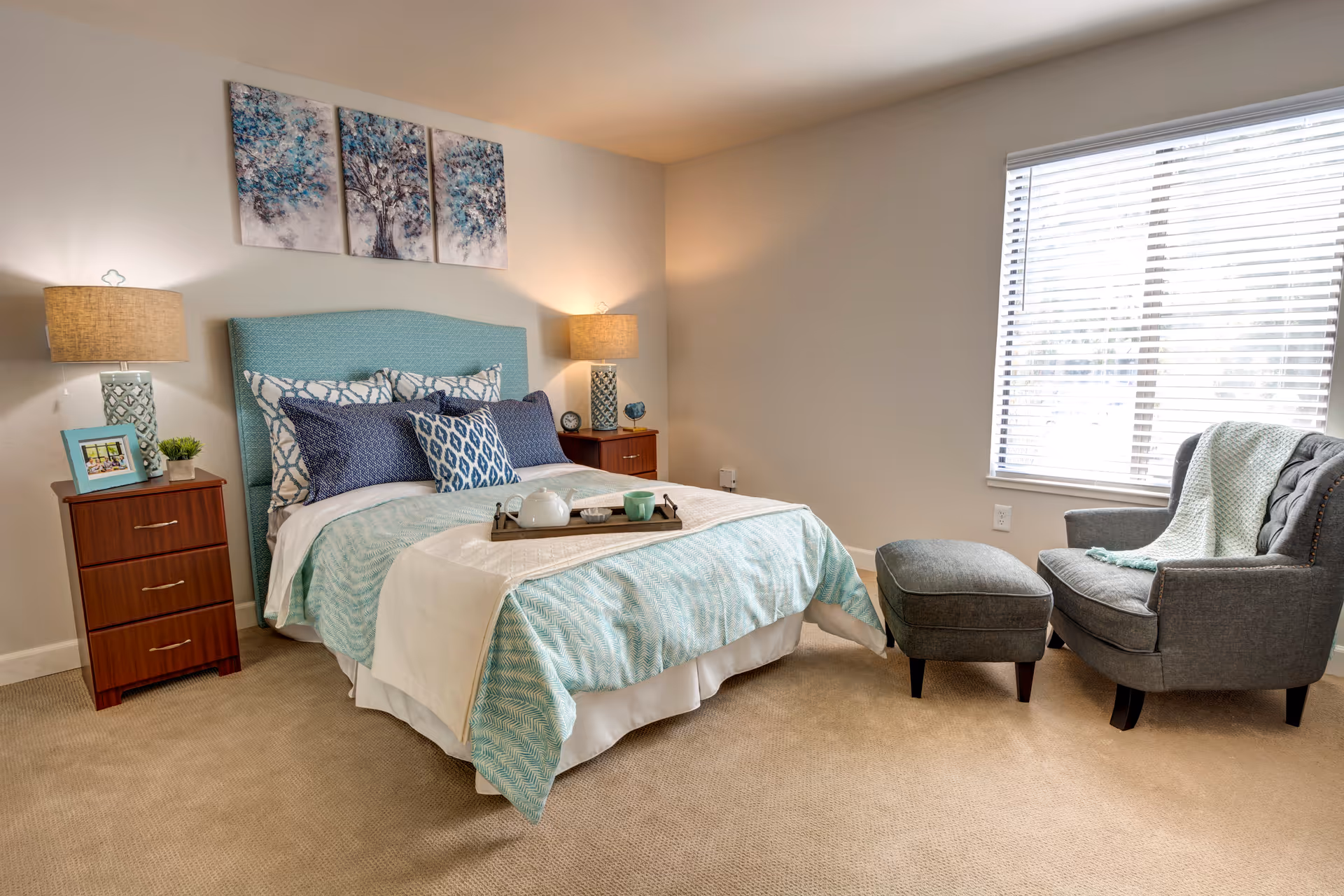 A cozy bedroom with a bed featuring a teal upholstered headboard, multiple patterned pillows, and a light blue and white bedspread. There are two wooden nightstands on either side of the bed, each with a lamp and decorative items. A gray armchair with a matching ottoman and a light blue throw blanket is positioned near a window with white blinds, allowing natural light into the room. Above the bed, there are three pieces of artwork depicting abstract trees in blue tones.