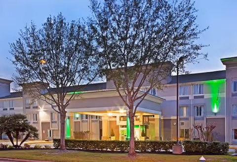 Exterior view of the Tunnel to Towers Veterans Village building at dusk, showing a three-story structure with illuminated green lights on the facade, a covered entrance with glass walls, trees, and landscaping in the foreground.