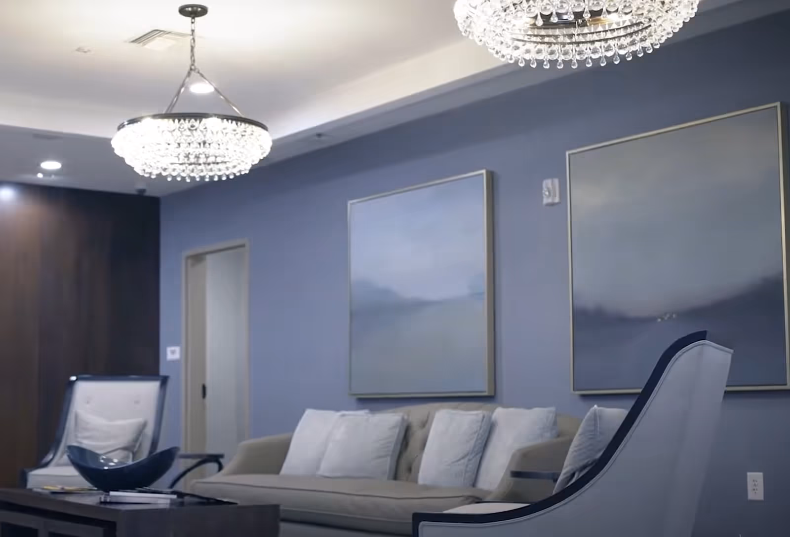 Stylish lounge with a sofa, armchairs, crystal chandeliers, and large abstract paintings on a blue-gray wall.