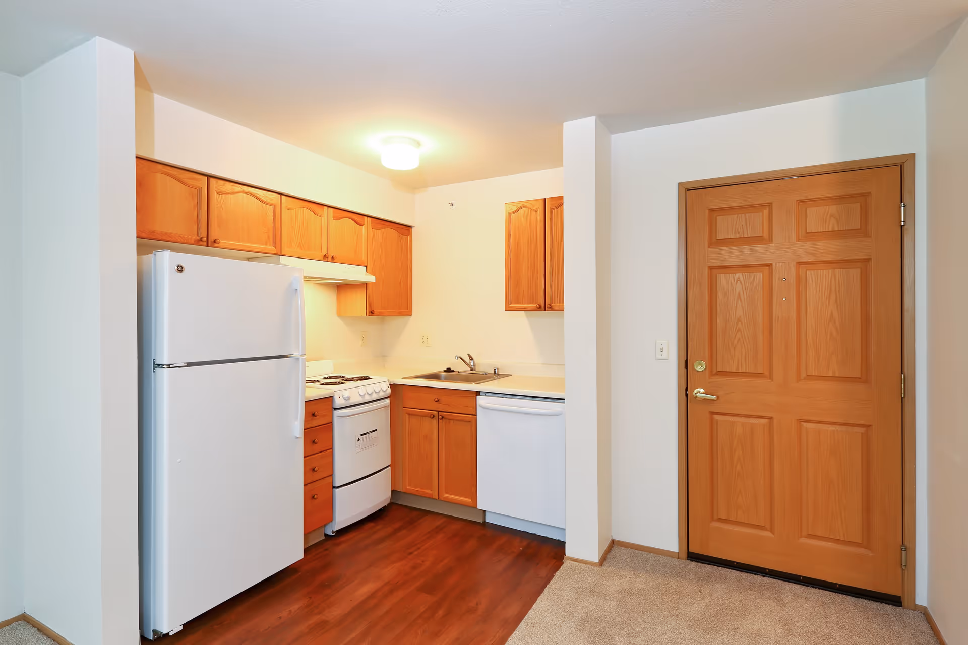 Small kitchen area with wooden cabinets, white refrigerator, white stove, white dishwasher, and a wooden entrance door. The floor is a combination of wood and carpet.