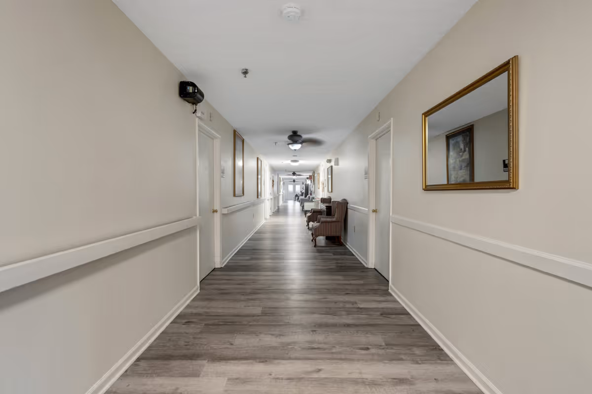 A long, well-lit hallway in a senior living facility with light-colored walls, wood flooring, and several closed doors on both sides. There are framed pictures and mirrors on the walls, ceiling fans, and a few upholstered chairs placed along the right side of the hallway.