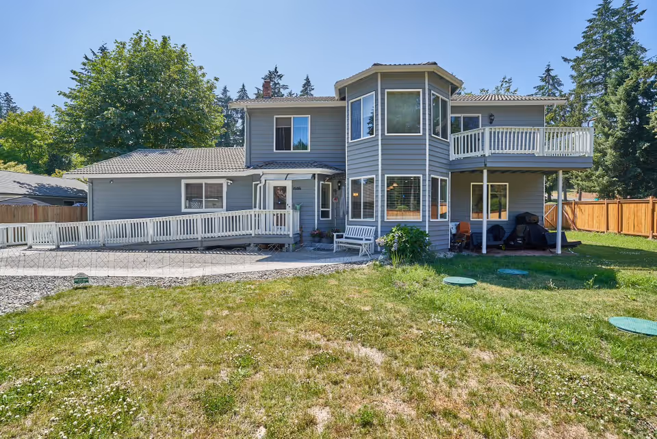 Exterior view of a two-story gray house with white trim, featuring a wheelchair ramp, a small porch with a bench, a balcony on the second floor, and a grassy yard with some trees and a wooden fence in the background under a clear blue sky.