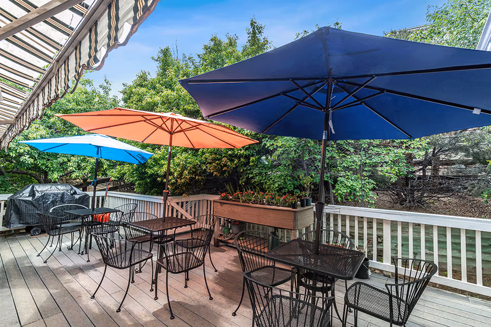 A wooden deck patio with black metal tables and chairs under blue, orange, and teal umbrellas beside a striped awning and planter boxes, overlooking trees.