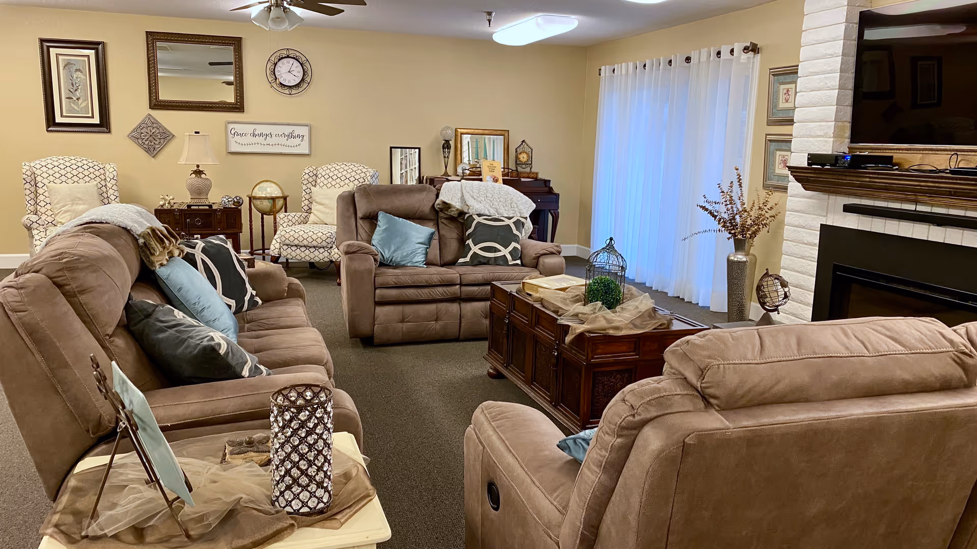 A cozy living room with beige walls and carpet, featuring three brown recliner sofas with various decorative pillows and throws. A wooden coffee table with decorative items sits in the center. The room has framed artwork and a clock on the wall, a ceiling fan, a white brick fireplace with a TV mounted above it, and sheer white curtains covering a window or door.