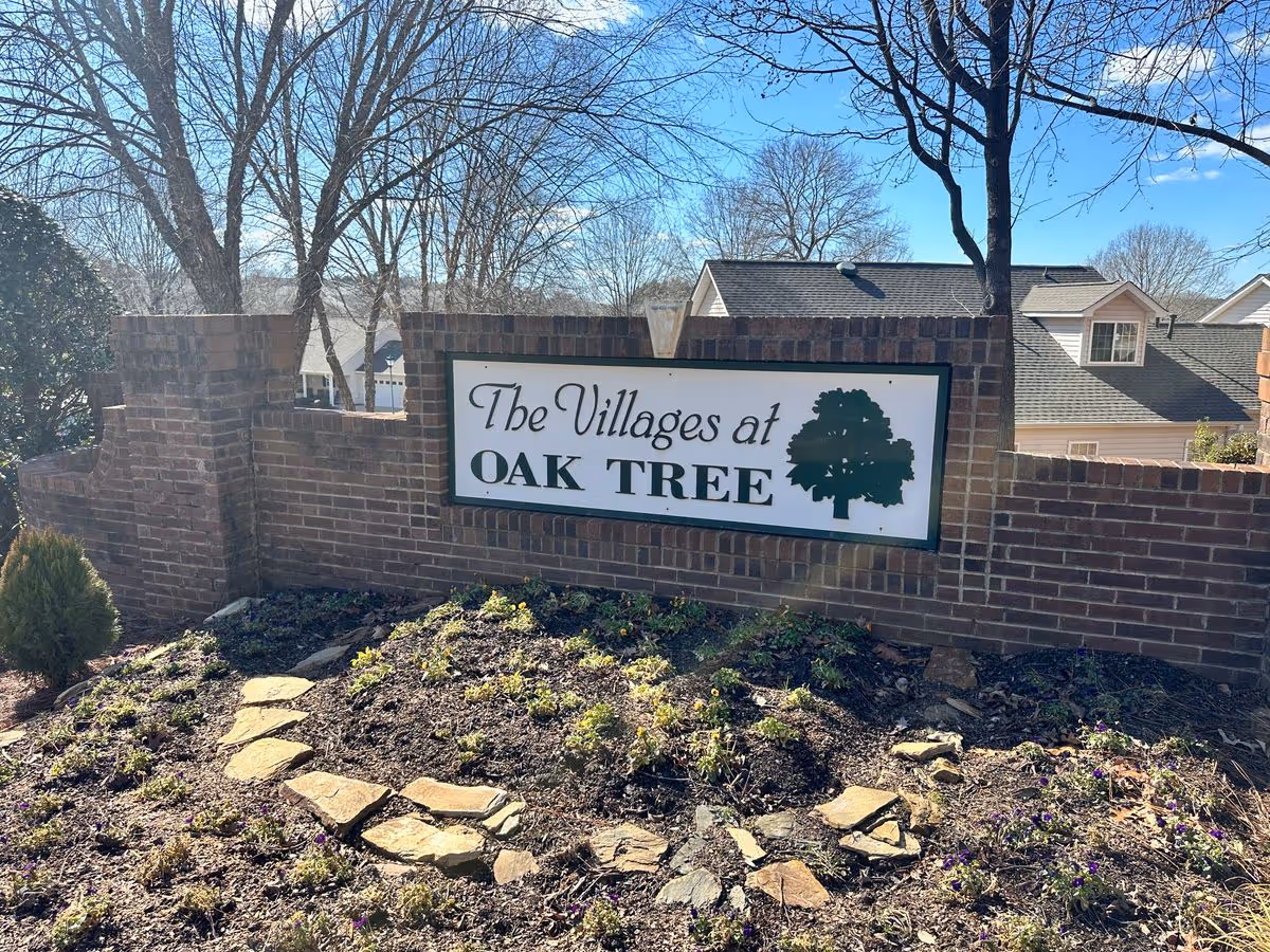 Brick entrance sign that reads 'The Villages at Oak Tree' set in a landscaped bed with houses and trees behind.