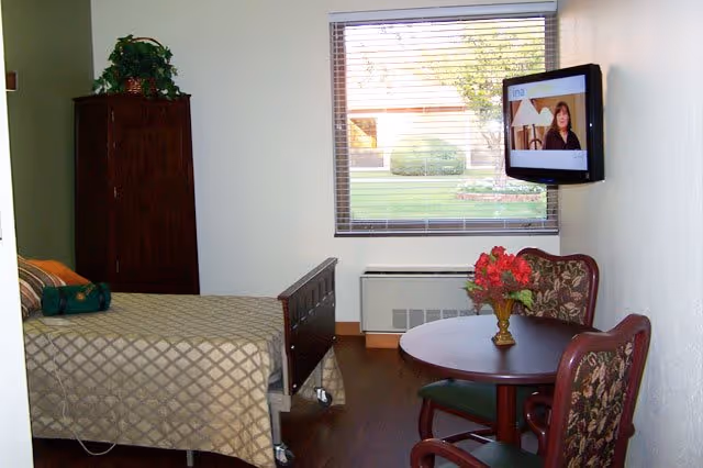 Small furnished senior living bedroom with a bed, wall-mounted TV, window, and a round table with two chairs.
