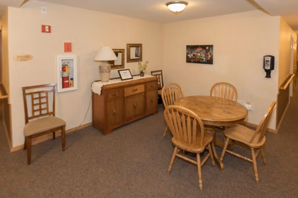 Small communal dining area with a round wooden table and chairs, a sideboard with lamp and framed art on the wall.