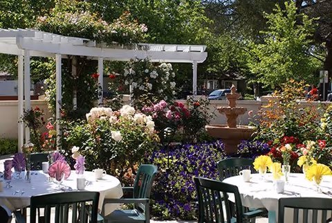 Outdoor patio area with round tables covered in white tablecloths, each set with cups, napkins folded in decorative shapes, and chairs. The patio is surrounded by lush flowering plants and greenery, with a white pergola and a multi-tiered stone fountain in the background.