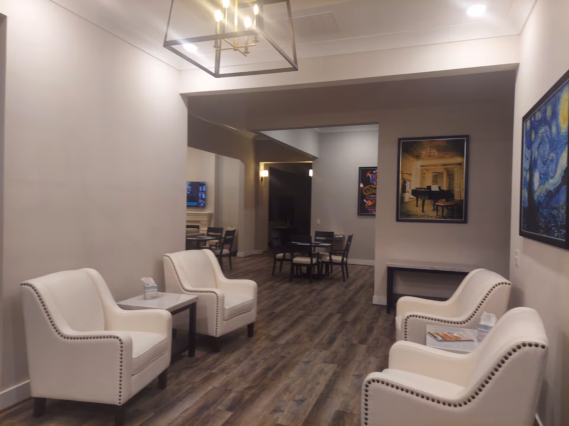 Bright common lounge with four white upholstered armchairs, side tables, wood-look flooring, and a dining area visible in the background.