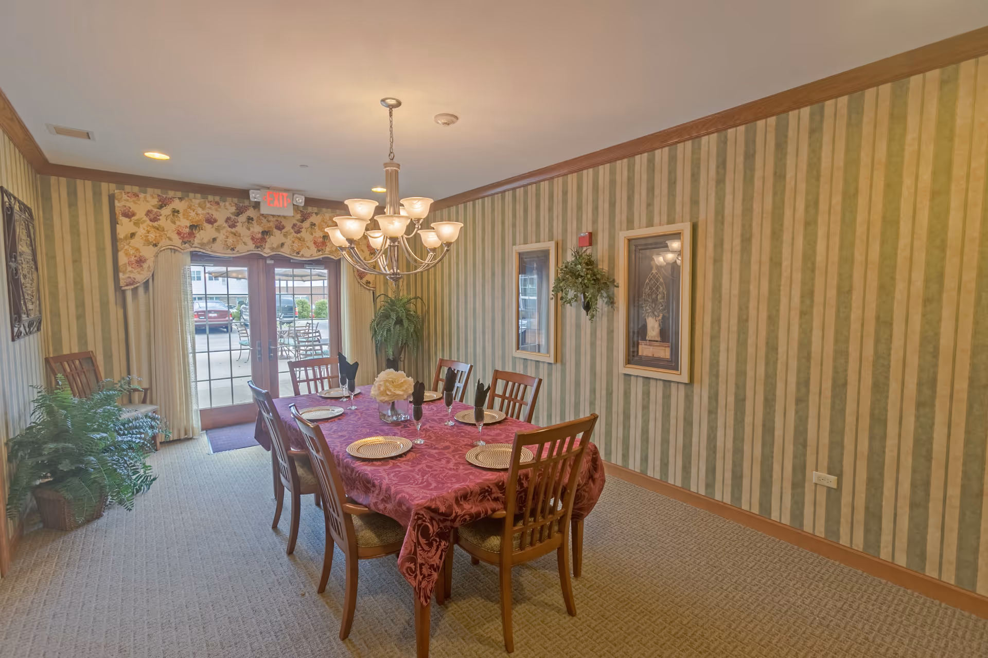 A dining room with a rectangular table covered with a red tablecloth set with plates, glasses, and napkins. Six wooden chairs surround the table. The room has green and beige striped wallpaper, two framed pictures, a hanging plant, and a chandelier overhead. French doors with floral curtains lead outside to a patio area with outdoor furniture.
