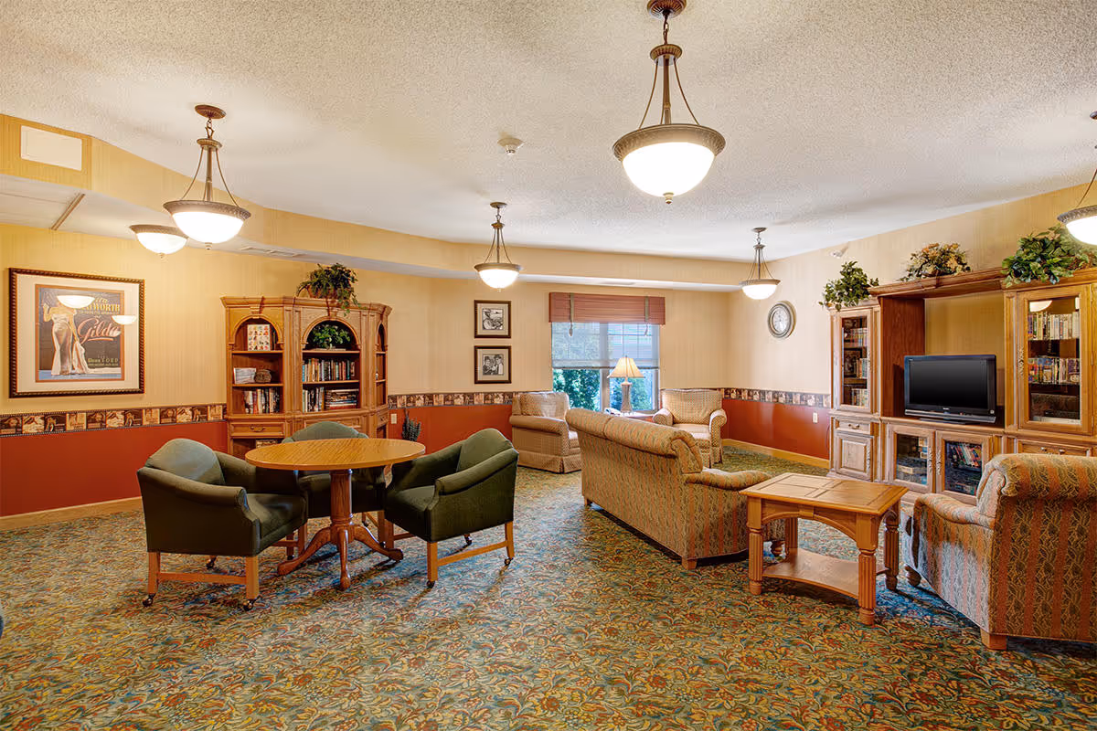 Spacious senior living lounge with sofas, armchairs, a round table, bookshelves and a TV cabinet under pendant lights.