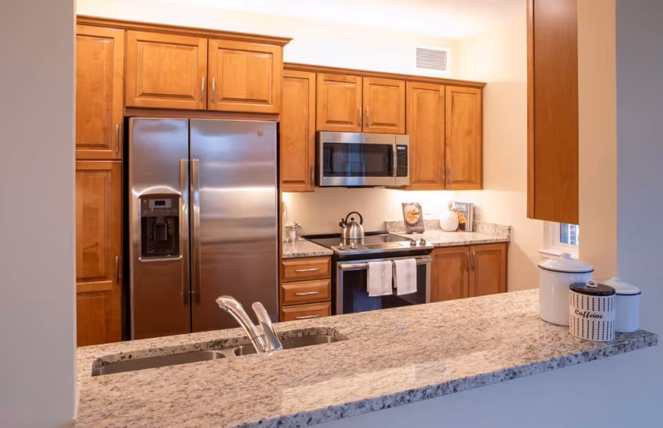 Modern kitchen with wooden cabinets, stainless steel refrigerator, microwave, and stove. Granite countertop with a double sink and containers labeled 'Caffeine' and others are visible.
