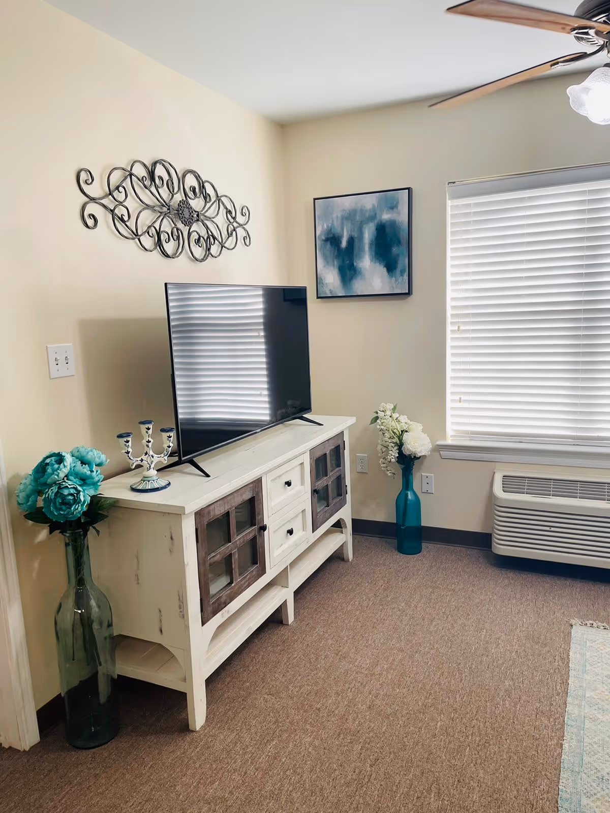 A cozy living room corner with a white wooden TV stand holding a flat-screen television. On the TV stand, there is a decorative candle holder. To the left of the stand is a tall glass vase with blue flowers, and to the right is another tall vase with white flowers. A window with closed blinds and an air conditioning unit below it is on the right wall. Above the TV stand, there is a decorative metal wall art and a framed abstract painting with blue and white tones. The room has beige walls, a ceiling fan with lights, and carpeted flooring.