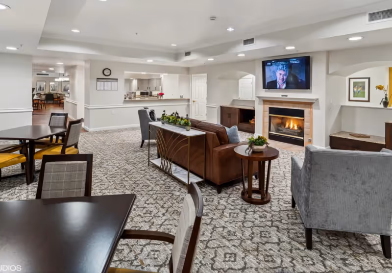 Open carpeted common area with seating, tables, a fireplace and a wall-mounted TV.