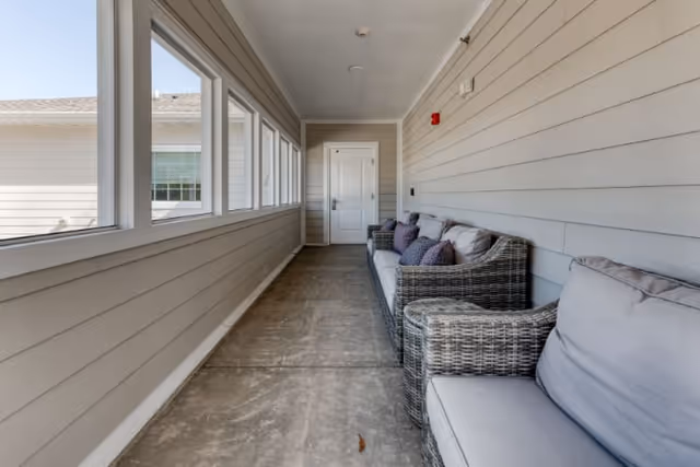 A long, narrow indoor hallway with large windows on one side and a row of cushioned wicker chairs with pillows on the other side. The walls are painted light gray with horizontal paneling, and there is a white door at the end of the hallway.