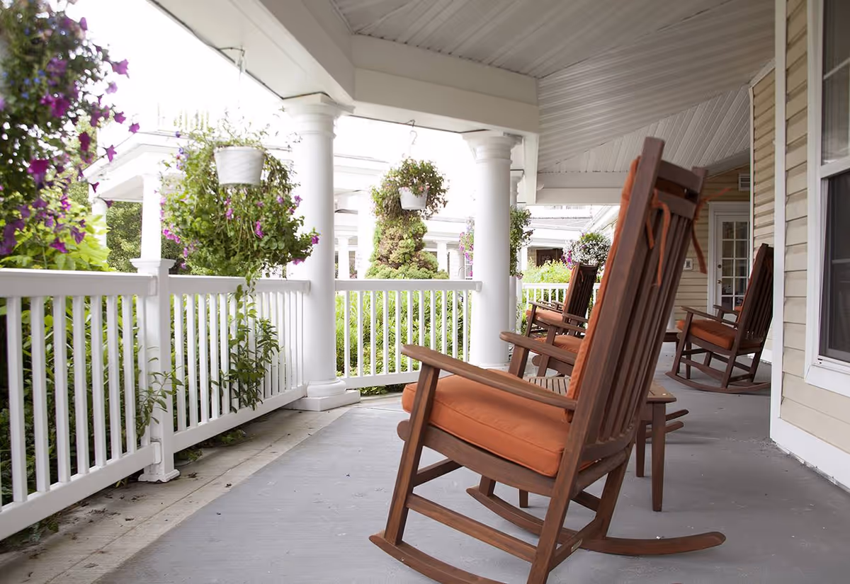 A covered porch area with wooden rocking chairs featuring orange cushions. The porch has white railings and columns, with hanging flower pots and lush greenery surrounding the space.