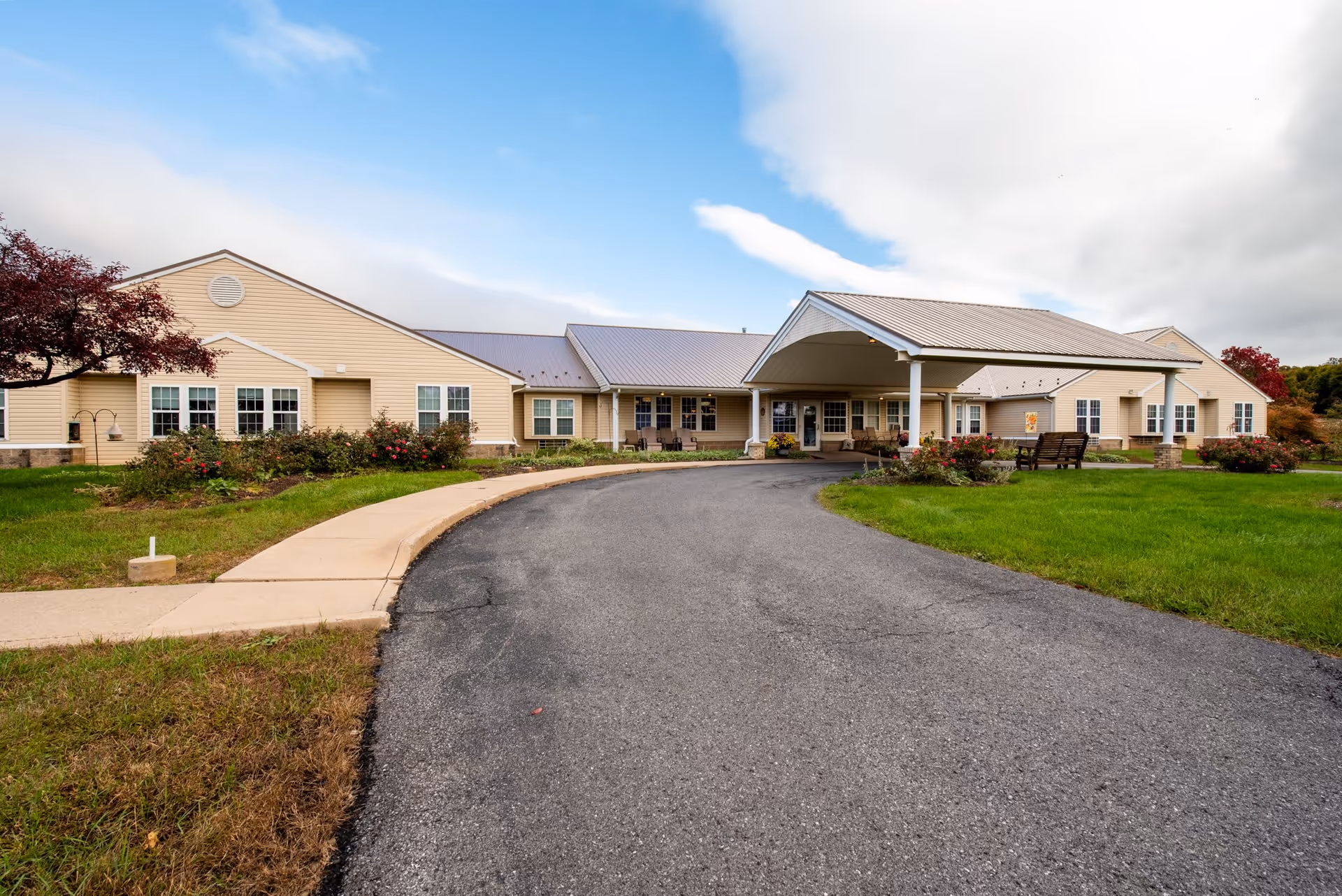 Single-story beige residential building with a covered porte-cochère, curved driveway, and landscaped lawn under a partly cloudy sky.