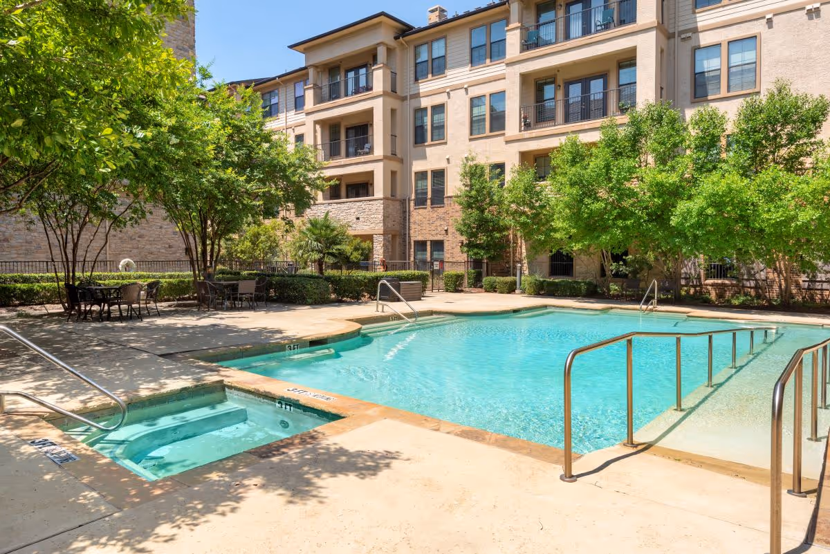 Outdoor swimming pool and adjacent hot tub with patio seating beside a multi-story senior living building and landscaped trees.