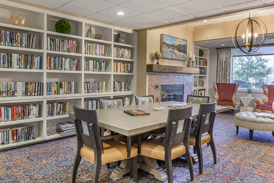 Cozy common room with a large table and chairs, built-in bookshelves, a fireplace, and seating near a window.