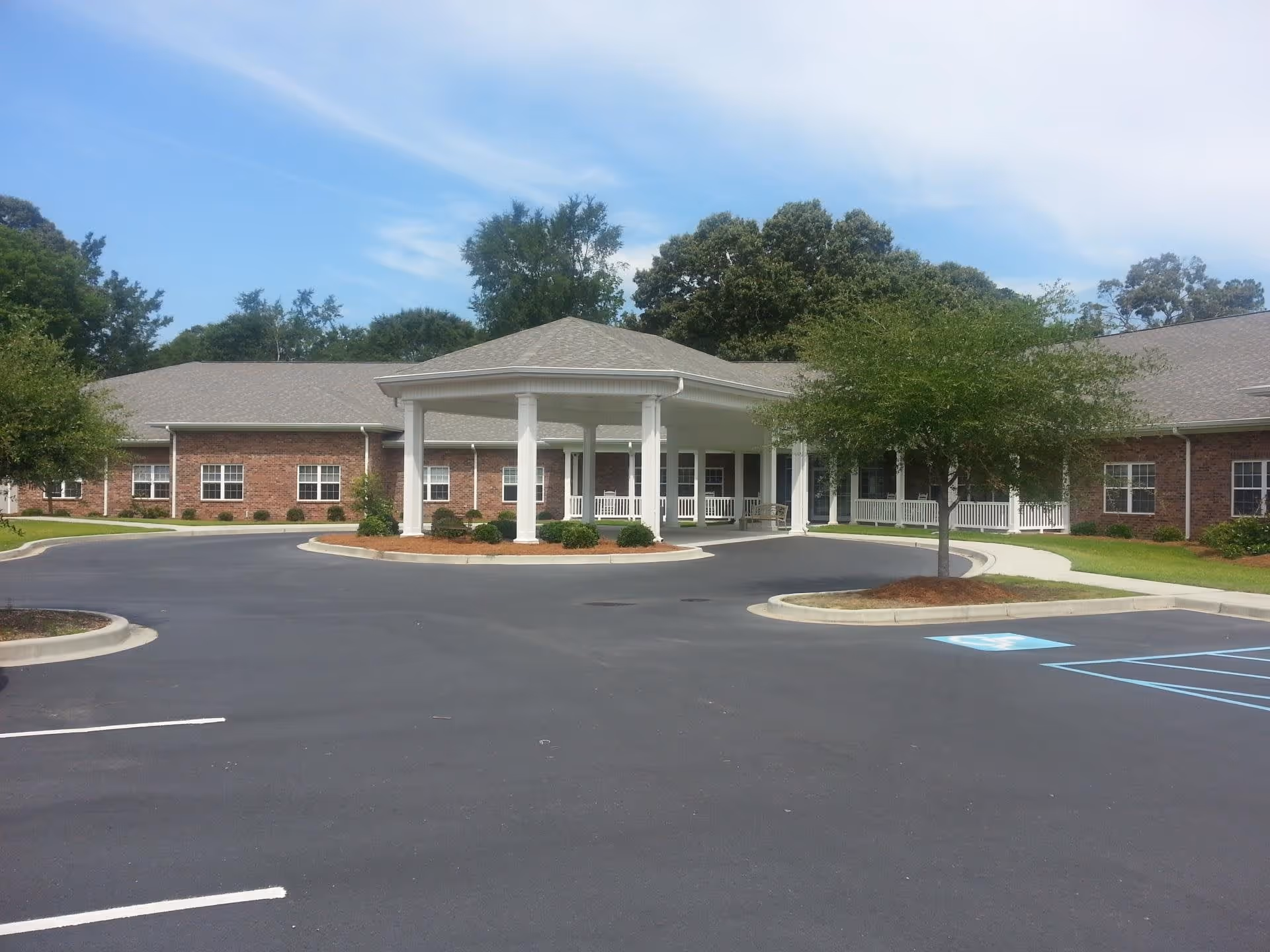 Front exterior view of a single-story brick building with a covered entrance supported by white columns, surrounded by a paved driveway and parking area with a handicapped parking space, some trees, and a partly cloudy sky.