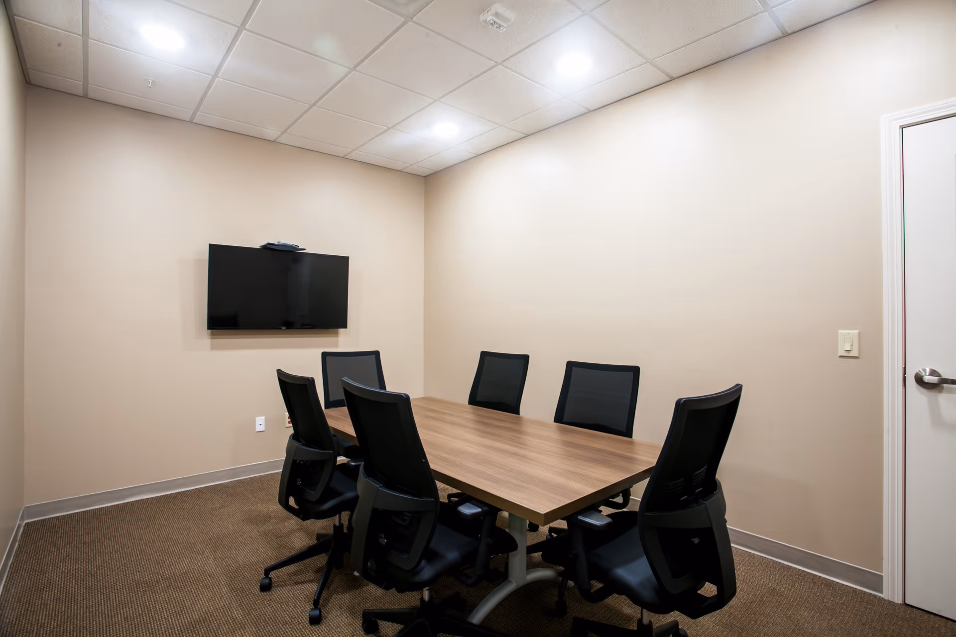 Small meeting room with a rectangular wooden table, six black office chairs, and a wall-mounted TV on a beige wall.