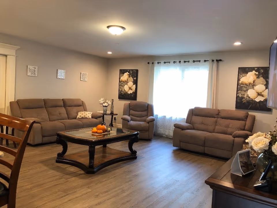A cozy living room with two beige sofas, a matching armchair, a wooden coffee table with decorative pumpkins, and floral artwork on the walls. The room has wood flooring, a window with sheer curtains, and soft ceiling lighting.