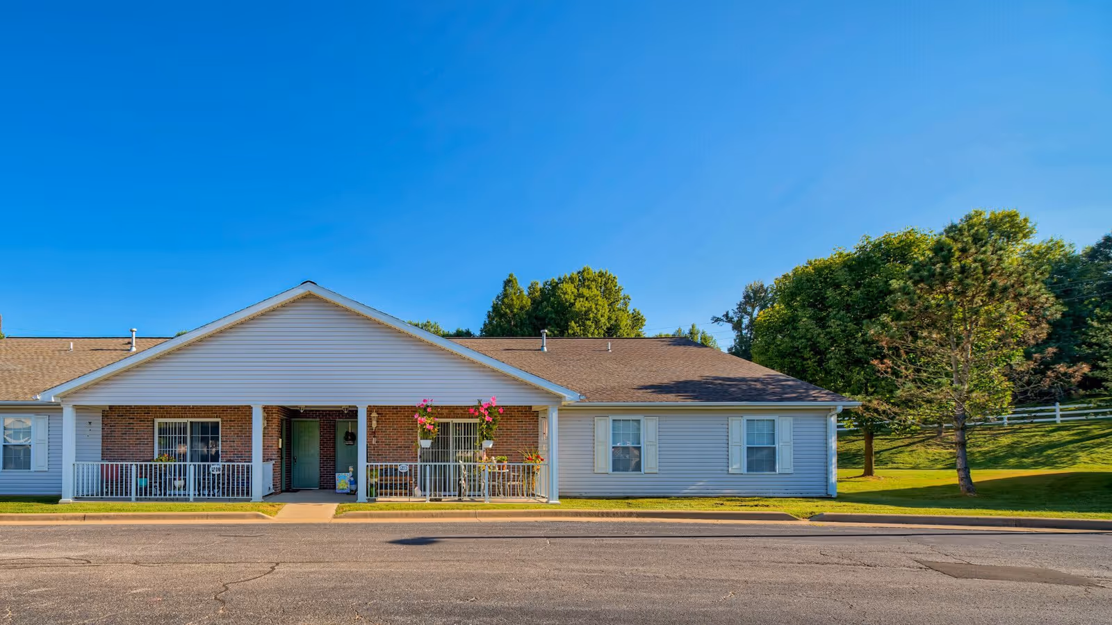 Single-story residential building with a covered porch featuring white railings and hanging flower pots, surrounded by green grass and trees under a clear blue sky.