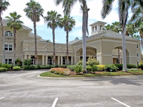 Front exterior of a large beige senior living building with a porte-cochere, palm trees, and a landscaped circular driveway.