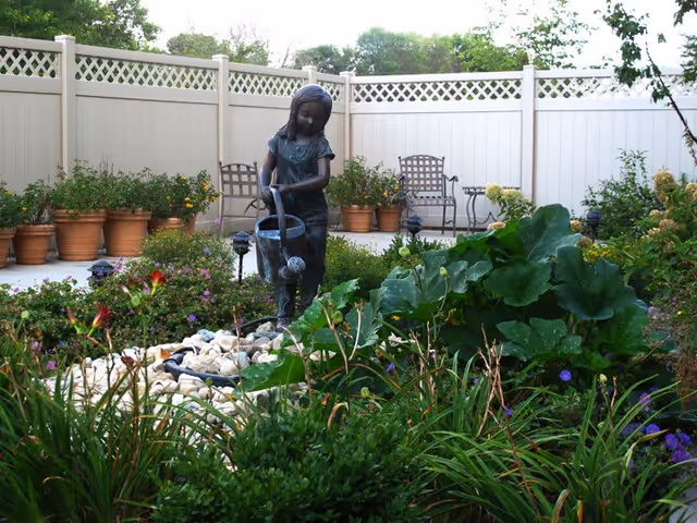 Small courtyard garden with a bronze statue of a girl pouring water, surrounded by plants, potted flowers, and patio chairs against a white fence.