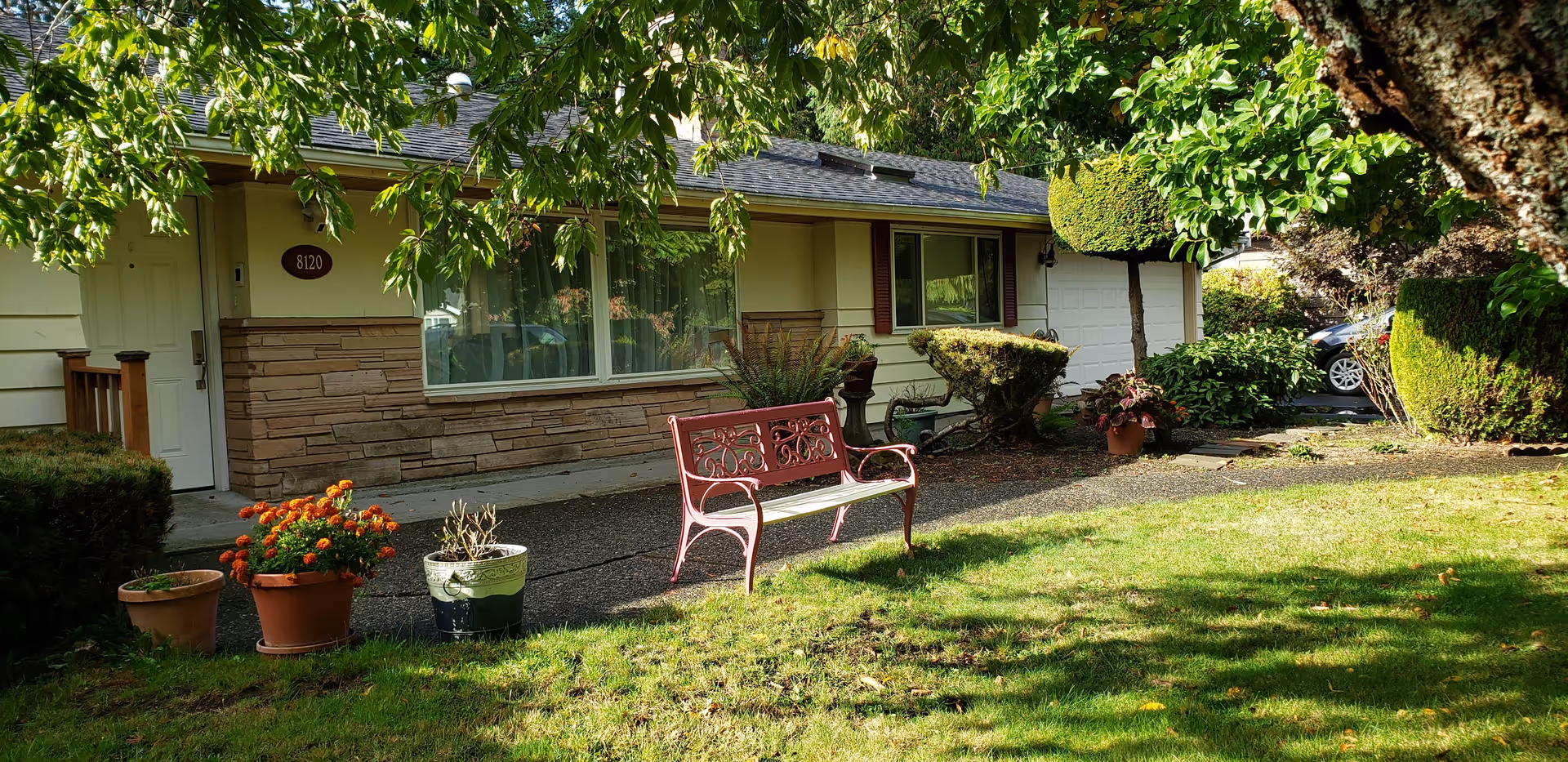 Front exterior view of a single-story house with a white door, large window, and attached garage. The house is surrounded by green grass, trees, and various potted plants. A red metal bench is placed on the concrete pathway in front of the house.