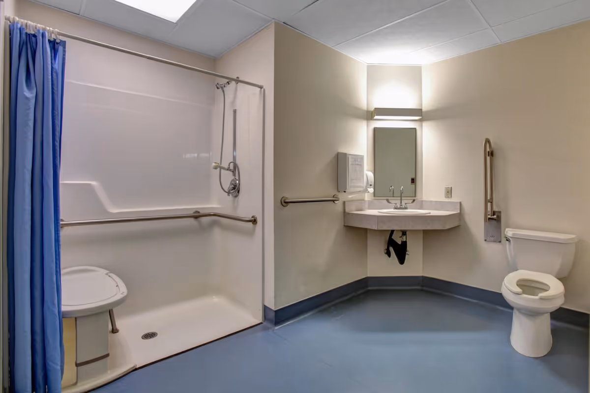 A spacious, accessible bathroom with a walk-in shower featuring a blue curtain and a built-in shower seat. The bathroom also includes a wall-mounted sink with a mirror and light above it, and a toilet with a raised seat and grab bars for support. The floor is blue, and the walls are light-colored.