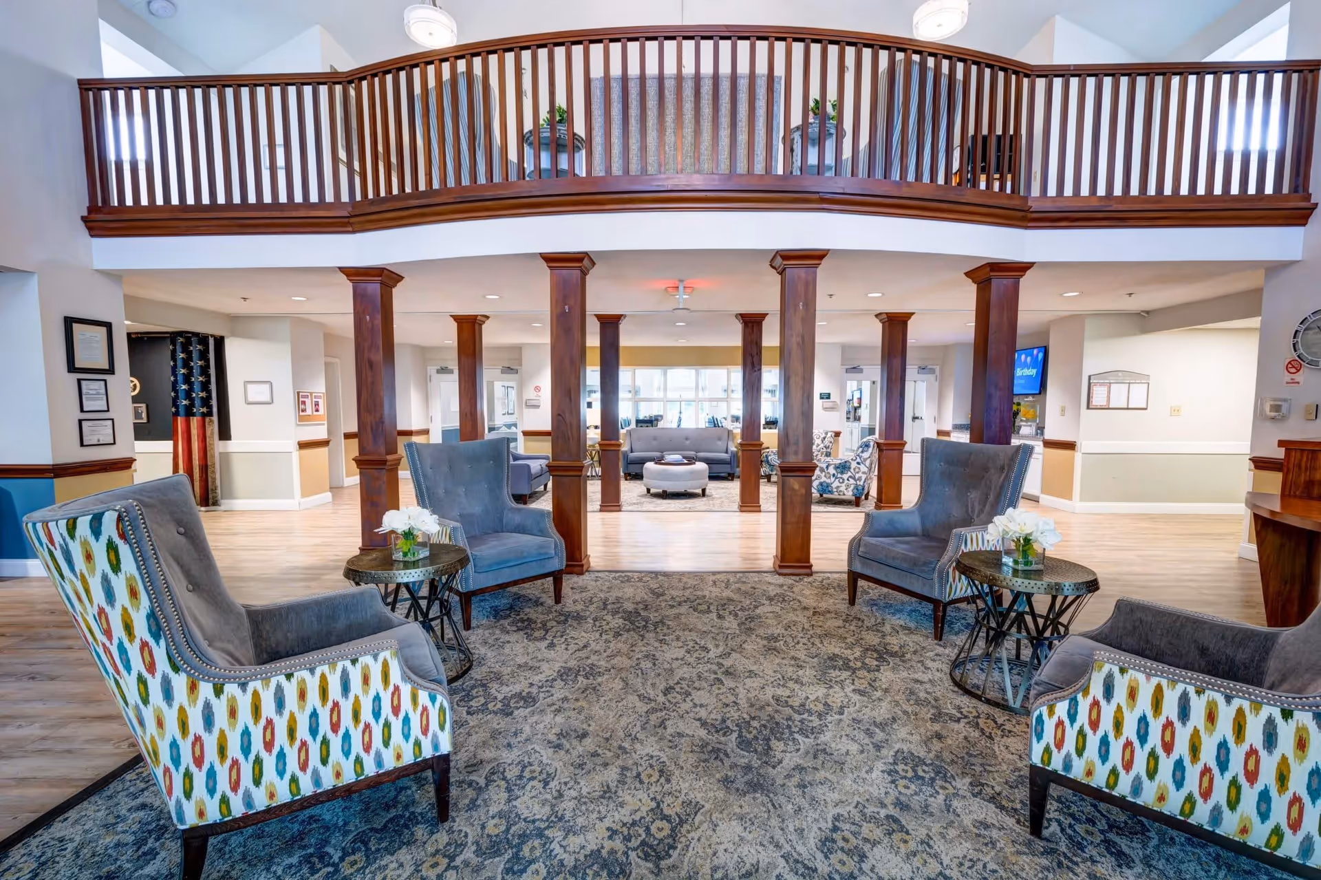 A spacious and well-lit senior living facility common area with a high ceiling and wooden balcony railing above. The room features multiple comfortable armchairs with colorful patterned upholstery arranged around small round tables with flower vases. Wooden columns separate the seating areas, and additional seating with sofas and chairs is visible in the background. The floor is covered with a large patterned rug and light wood flooring.