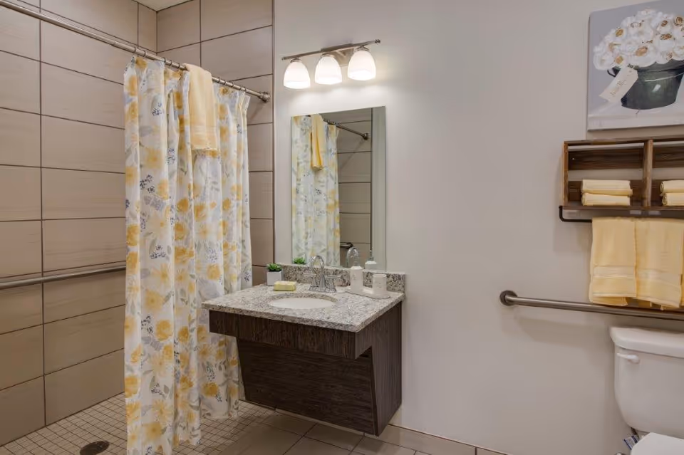 A clean and modern bathroom featuring a floral-patterned shower curtain with yellow and gray flowers, a wall-mounted granite countertop sink with a mirror above it, three light fixtures, and a wooden shelf holding folded yellow towels above a toilet. There are grab bars installed near the shower and toilet for accessibility.