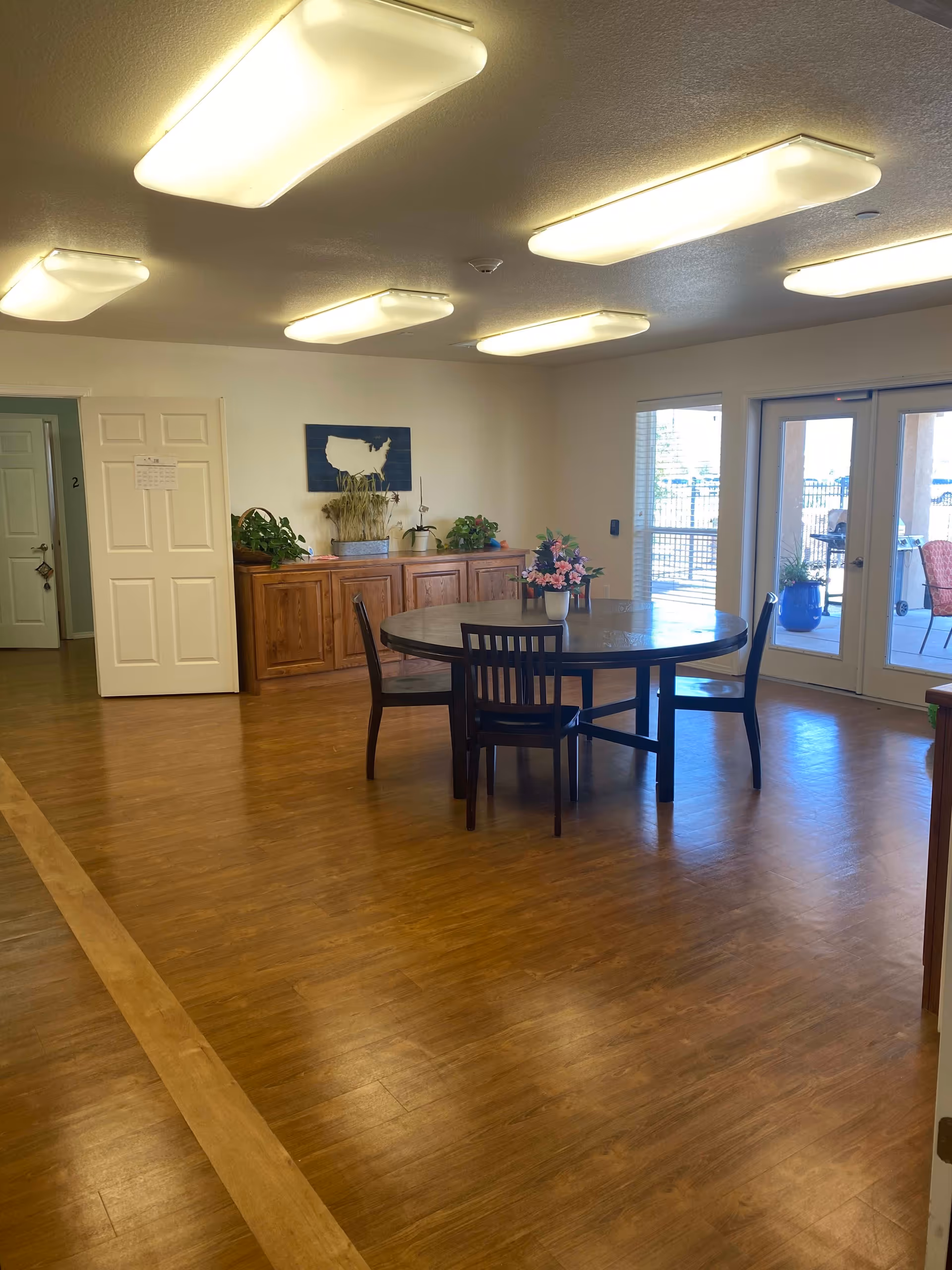 A spacious room with wooden flooring featuring a round wooden table with four chairs around it. On the table, there is a vase with pink flowers. Against the far wall, there is a wooden cabinet with several potted plants and a decorative wall art piece shaped like the United States. To the right, there are glass double doors leading to an outdoor area with patio furniture and plants. The ceiling has multiple rectangular fluorescent light fixtures.