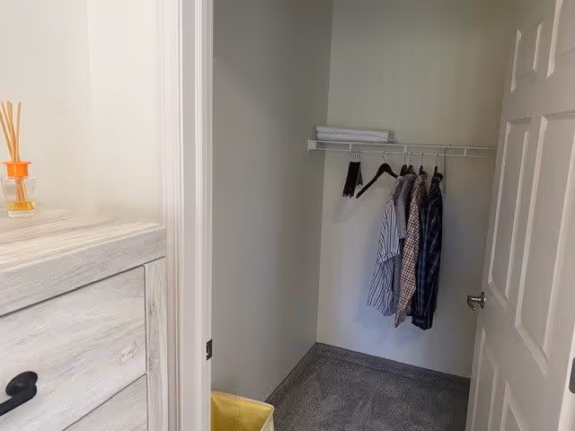 A small closet with a hanging rod holding four shirts on hangers and a shelf above with folded towels. To the left of the closet is a light wood dresser with a small reed diffuser on top. The closet door is open, showing a carpeted floor and a yellow laundry basket on the floor.