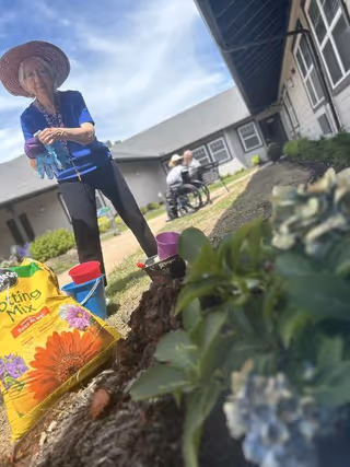 An elderly woman wearing a wide-brimmed hat and blue shirt is gardening outdoors near a building. There are gardening supplies including a bag of potting mix and colorful buckets on the ground. In the background, an elderly man in a wheelchair is visible near the building under a partly cloudy sky.