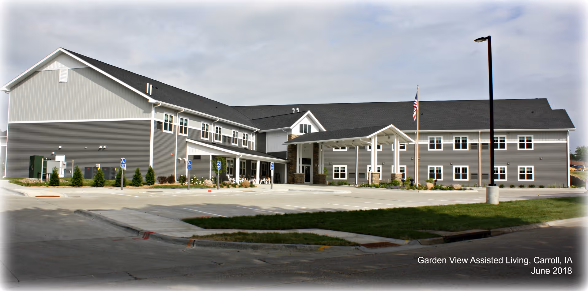 Exterior view of Garden View Assisted Living facility in Carroll, IA, showing a two-story building with gray siding, multiple windows, a covered entrance with white pillars, an American flag on a flagpole, and a parking area with handicap parking spaces.