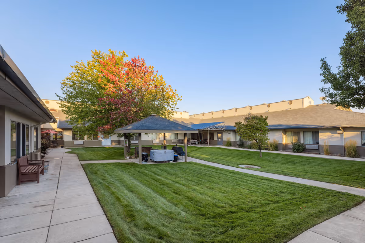 Outdoor courtyard area of a senior living facility with well-maintained green grass, a paved walkway, a gazebo with seating, and trees including one with autumn-colored leaves. The surrounding building is single-story with windows and doors facing the courtyard.
