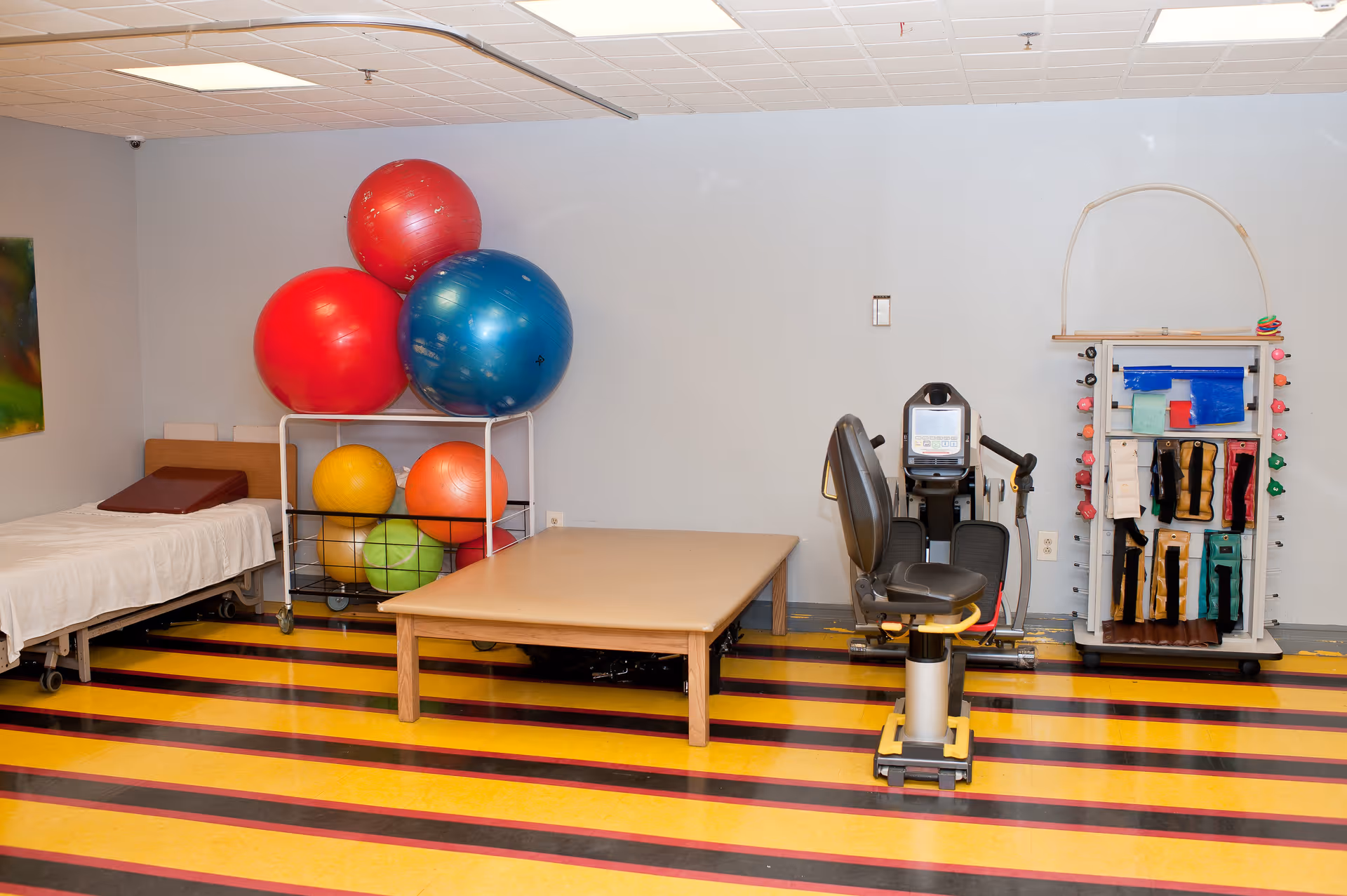 Physical therapy/exercise room with large colorful exercise balls, a treatment table, a recumbent exercise bike, and therapy equipment on a yellow-and-black striped floor.