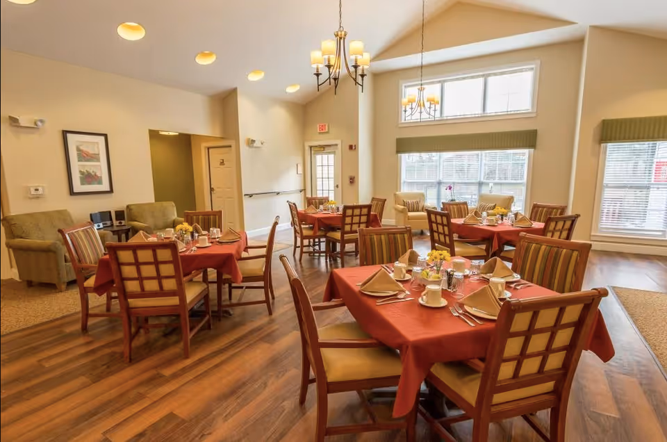 A bright dining room with several wooden tables covered with red tablecloths, each set with plates, cups, napkins, and silverware. The room has large windows with green valances, wooden flooring, and comfortable chairs. There are also a couple of armchairs and framed artwork on the walls.