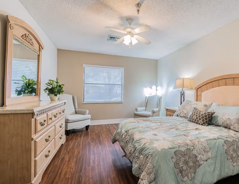 Furnished bedroom with a floral-patterned bed, dresser and mirror, two armchairs, a ceiling fan, and hardwood flooring.