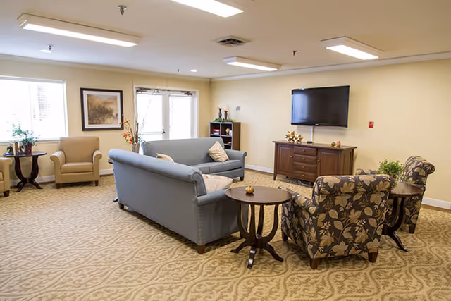 A cozy living room area in a senior living facility with a blue sofa, two patterned armchairs, a beige armchair, a flat-screen TV mounted on the wall above a wooden cabinet, and small round wooden tables. The room has beige walls, carpeted floor with a subtle pattern, and large windows letting in natural light.