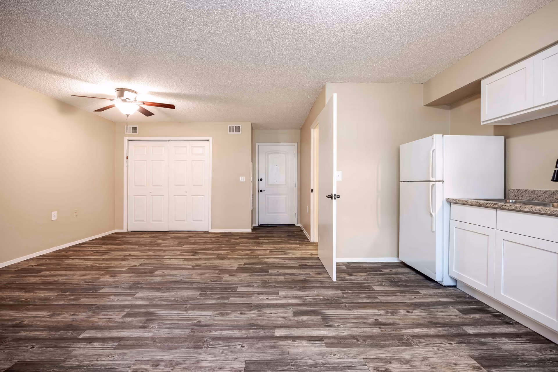 Empty room with wood-patterned flooring, beige walls, a ceiling fan with light, white double closet doors, a white entrance door, and a kitchen area with white cabinets, a countertop, and a white refrigerator.