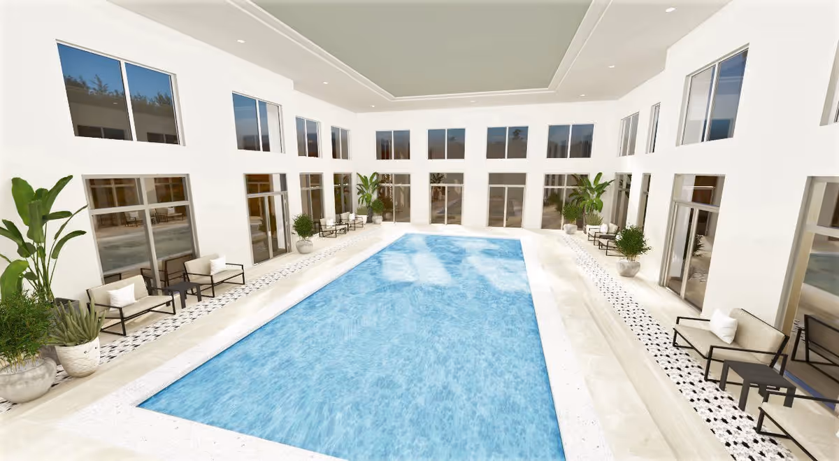 Indoor swimming pool area with clear blue water surrounded by white walls and large windows. There are several potted plants and seating areas with cushioned chairs and small tables along both sides of the pool.
