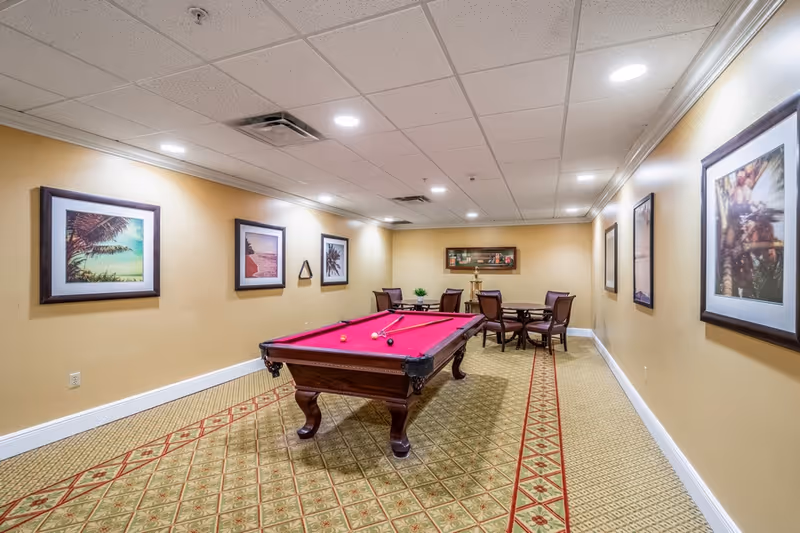 A recreational room with a red felt pool table in the center, surrounded by beige walls adorned with framed pictures. At the far end, there are two round tables with chairs arranged around them. The room has a patterned carpet and a drop ceiling with recessed lighting.