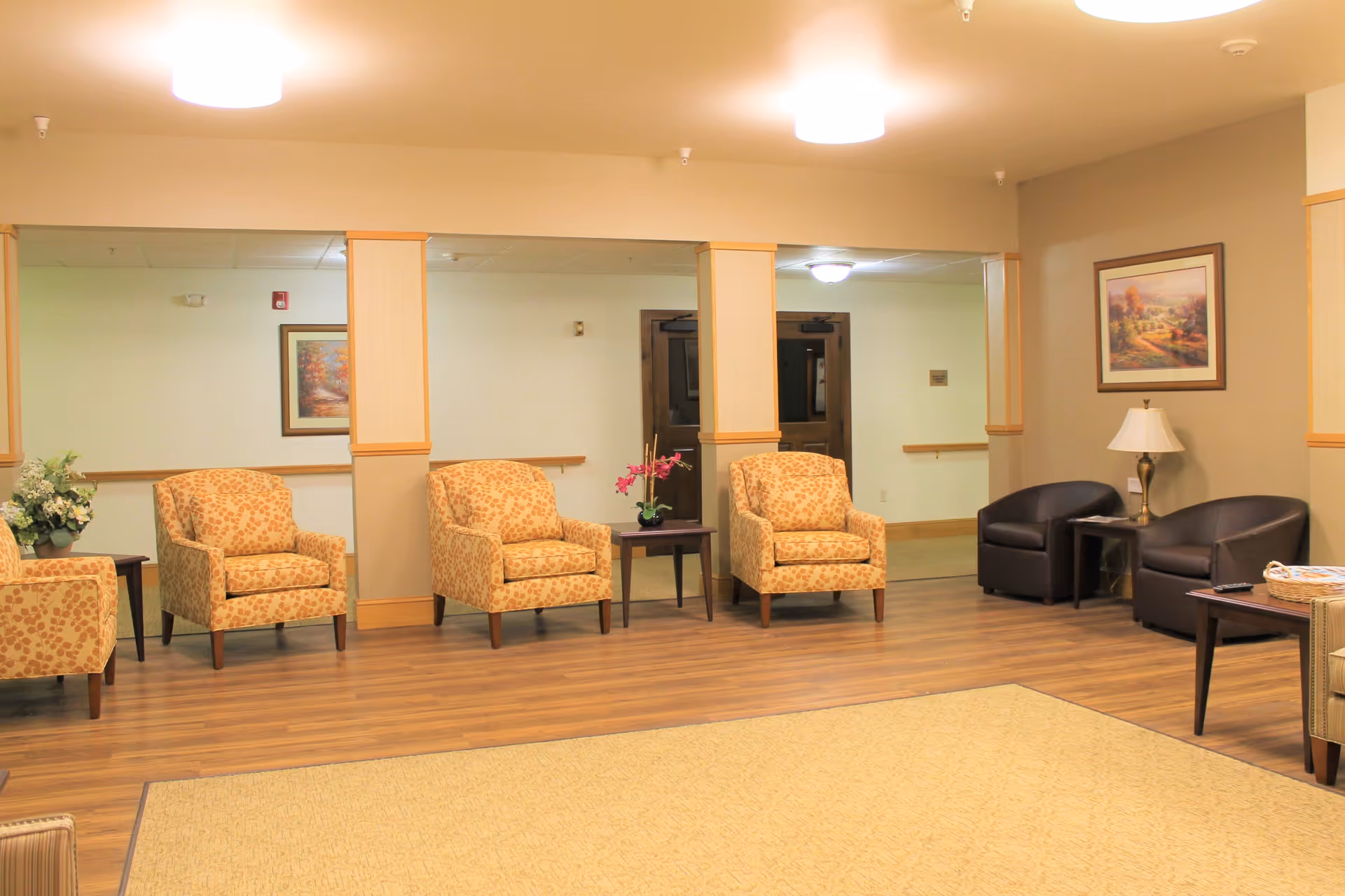 A spacious living room area with wooden flooring and beige walls. The room features several armchairs with patterned upholstery arranged along the walls, two dark leather chairs with a small table and lamp between them, framed artwork on the walls, and a beige carpet covering part of the floor. There are also small tables with decorative plants and flowers.