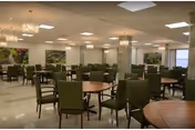 Large dining room with round wooden tables and green upholstered chairs beneath ceiling lights and chandeliers.